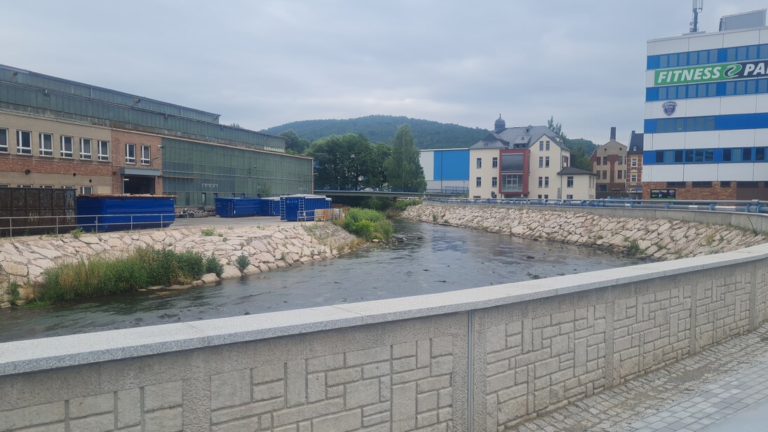 im Vordergrund eine niedrige Mauer, dahinter ein Fluss, der eine Kurve macht mit steinernem Ufer und angrenzender Bebauung