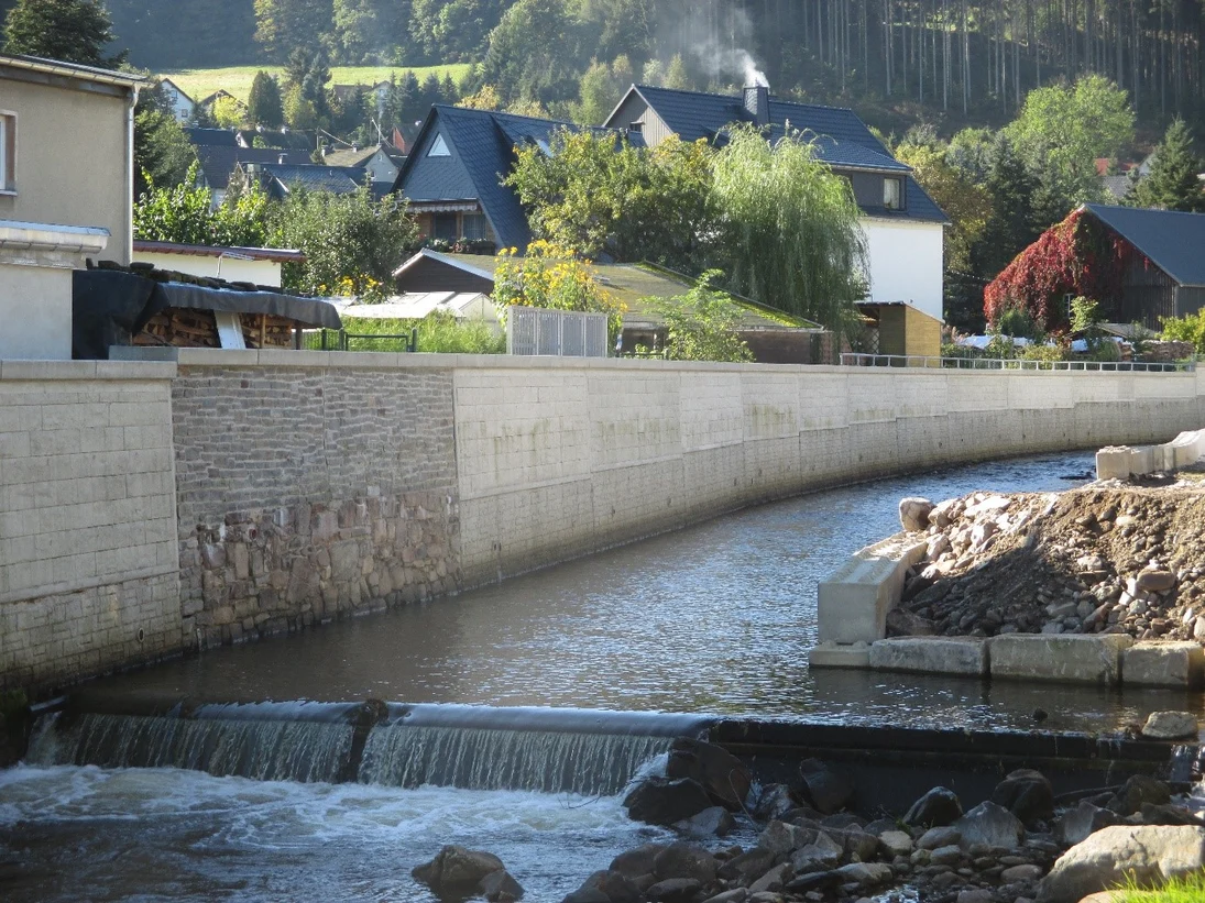 Fluss im Vordergrund mit angrenzender Steinmauer, dahinter Häuser