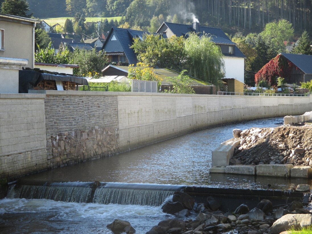 Fluss im Vordergrund mit angrenzender Steinmauer, dahinter Häuser