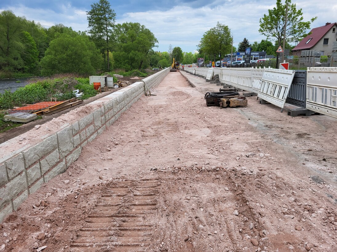 Blick in eine Baustelle mit kleiner Mauer links und Bäumen und Fluss im Hintergrund