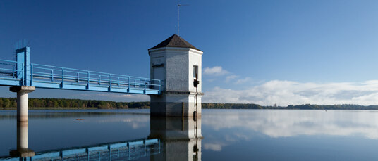 Blick auf einen blauen Metallsteg mit Türmchen am Ende, das in einen See ragt