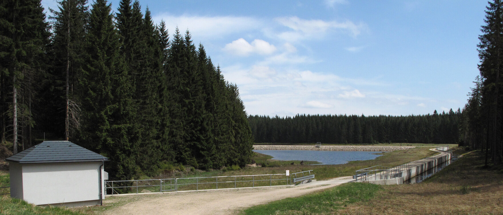 Blick auf einen Sandweg, links daneben steht ein kleines Gebäude vor einem Wald, am Ende des Weges befindet sich Wasser, darüber blauer Himmel