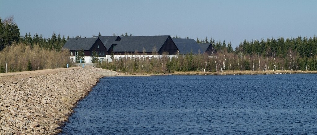 Blick auf einen See mit Steinufer auf der linken Seite, im Hintergrund Wald und Häuser