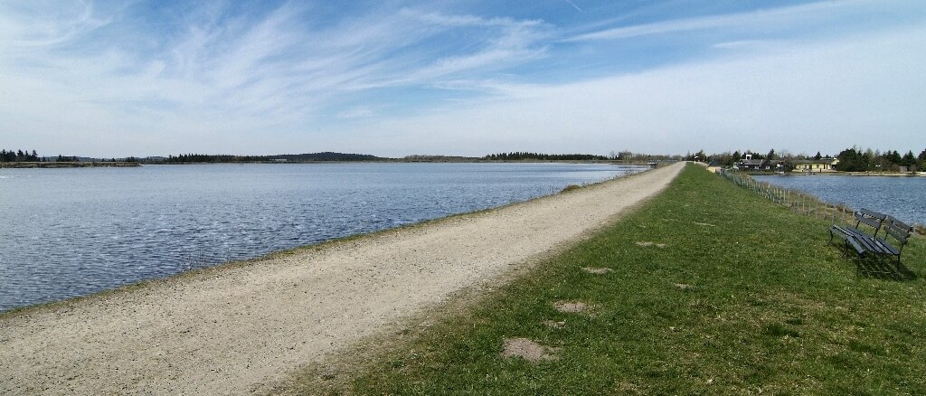 Blick vom Ufer mit Wiese auf einen See, in den ein glauer Metallsteg hineinragt.
