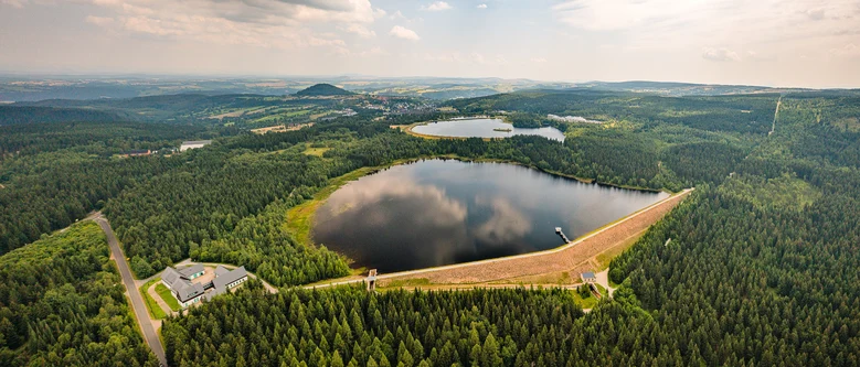 eine halbrunde Wasserfläche mit Staudamm, umgeben von viel Wald. Im Hintergrund eine zweite Wasserfläche