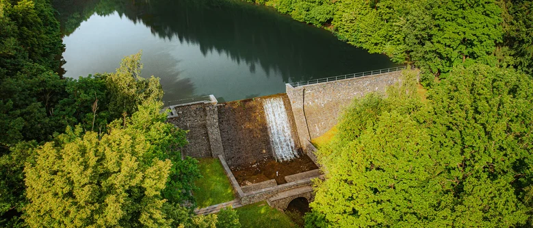 kleine Staumauer aus Stein mit Wasser dahinter, umgeben von grünen Bäumen