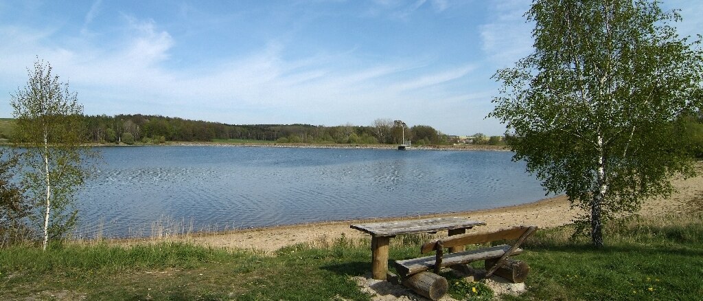 Blick vom Ufer auf einen See, am Ufer steht eine Holzbank mit einem Holztisch davor, rechts und links Bäume