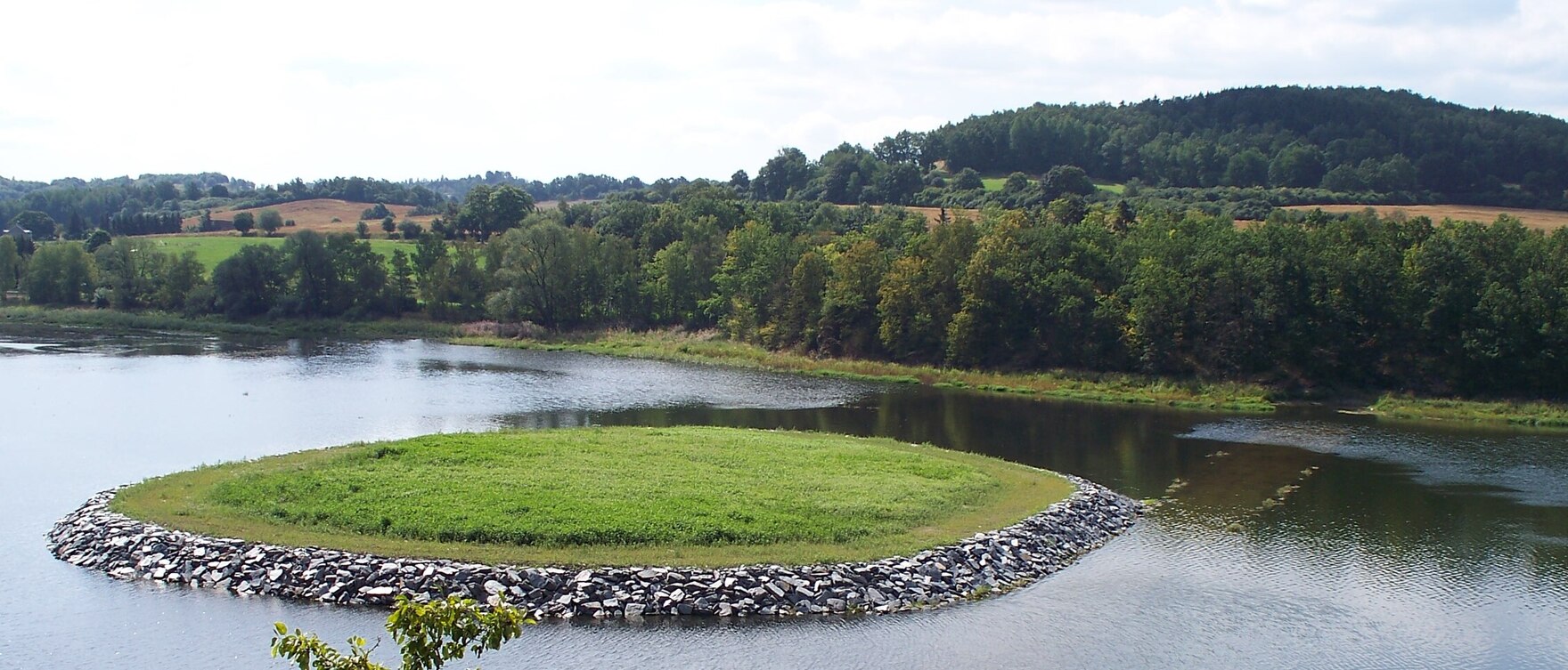 Blick auf einen See mit einer runden Wiese in der Mitte, die von Steinen begrenzt ist. Im Hintergrund Wald.
