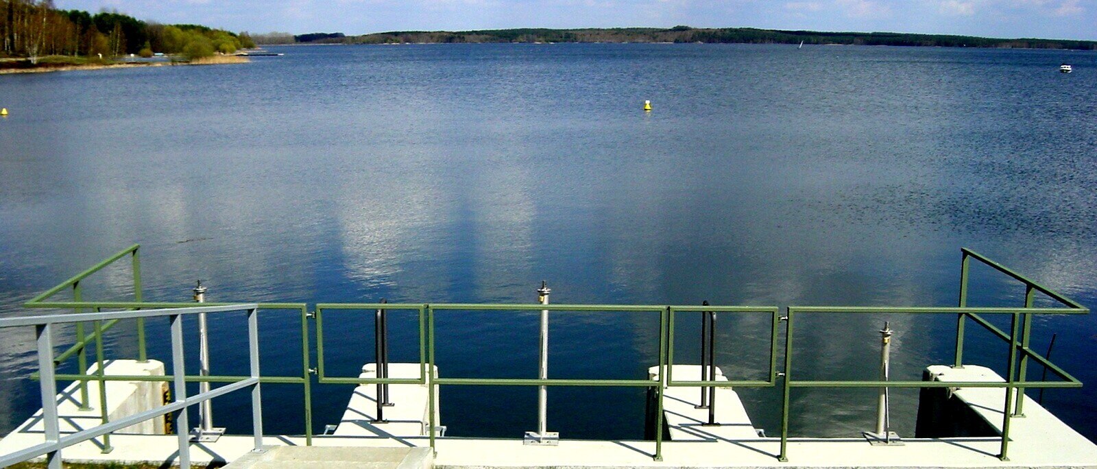 Blick auf einen See, am unteren Bildrand befindet sich ein Betonbauwerk mit grünem Metallgeländer
