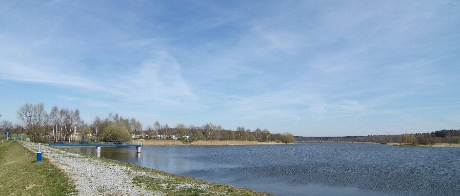 Blick vom Ufer auf einen See mit einem Weg im Vordergrund und Wald im Hintergrund und blauem Himmel