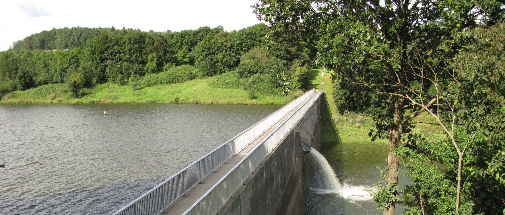 Blick vom erhöhten Ufer seitlich auf eine Staumauer. Rechts und links davon Wasser, im Hintergrund bewaldetes Ufer