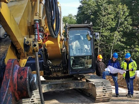 drei Bauarbeiter stehen an einem Raupenbagger und schauen in einen Plan