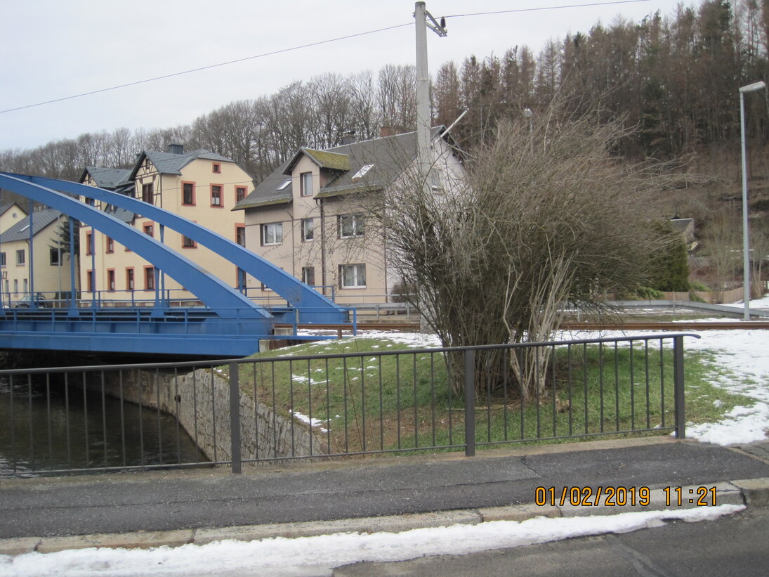 Blick von einer Brücke über einen Fluss auf eine blaue andere Brücke, dahinter Häuser