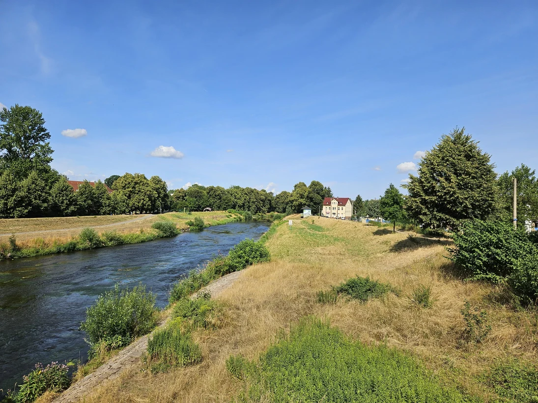 Fluss mit Uferwiesen und Bäumen unter blauem Himmel, im Hintergrund ein Haus