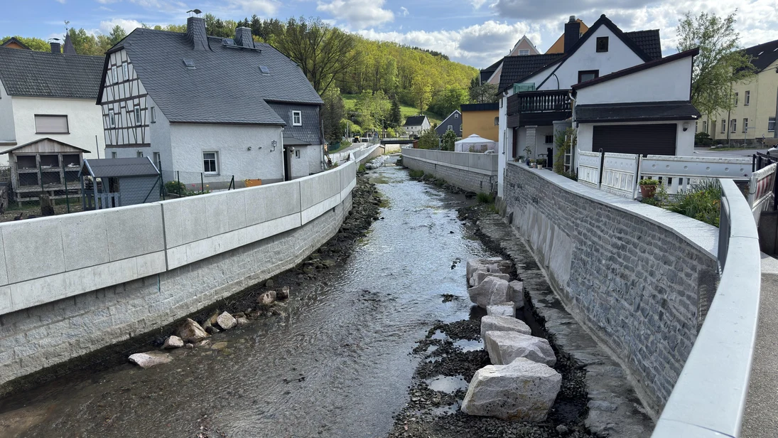 Blick auf einen Fluss mit neu errichteten Ufermauern aus Beton und Naturstein zwischen Wohnhäusern in einer ländlichen Umgebung