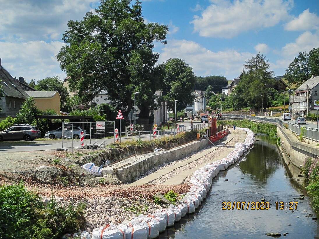 Blick auf einen Fluss an dessen rechter Seite eine Mauer verläuft und links Sandsäcke ihn in sein Bett zwingen, daneben eine Baustraße und ein Parkplatz